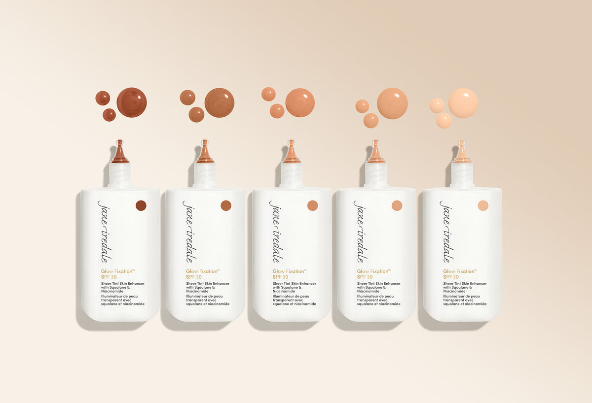 Fünf Flaschen des jane iredale Glow Fixation SPF 30 in verschiedenen Farbnuancen mit passenden Farbtropfen oberhalb der Verpackungen.