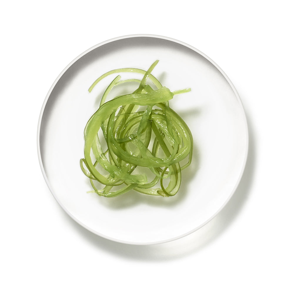 Draufsicht auf eine kleine weiße Schale mit grünen, spiralförmigen Algenstreifen auf weißem Hintergrund.
