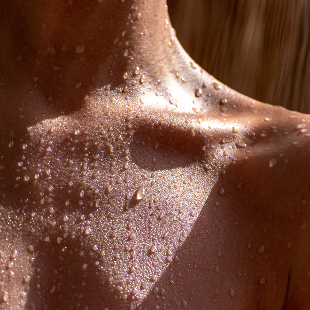 Detailaufnahme eines nackten oberen Brustbereichs mit glänzender, feuchter Haut, bedeckt von Wassertropfen. Das Licht betont die natürliche Textur und die gesunde Ausstrahlung der Haut.