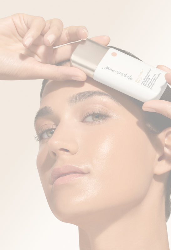 Nahaufnahme eines Models mit strahlender, natürlich wirkender Haut, das das jane iredale Glow Fixation SPF 30 über der Stirn präsentiert. Das Produkt wird mit einer Hand gehalten, während das Model selbstbewusst in die Kamera blickt. Der warme, weich verschwommene Hintergrund in Beige- und Sandtönen unterstreicht die hochwertige Beauty-Ästhetik und lässt ausreichend Freiraum für Text im linken Bildbereich.